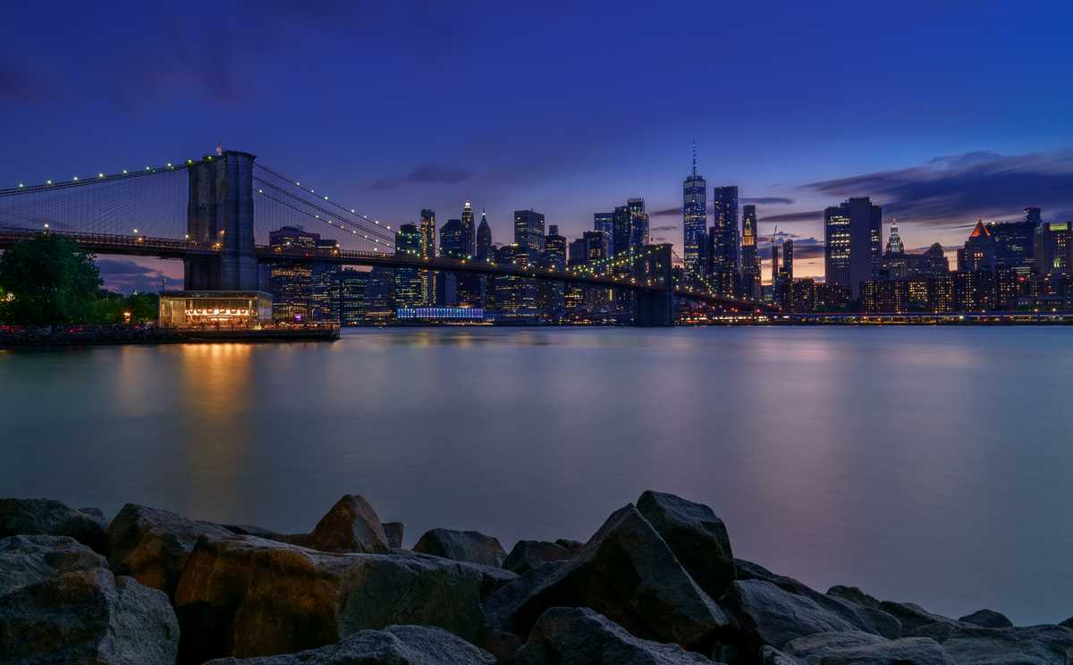 Brooklyn Bridge at twilight with the lit Manhattan skyline reflecting on the East River and rocks in the foreground